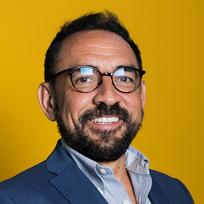 A man with glasses, a blue blazer, and a light blue collared shirt stands and smiles.