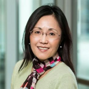 A woman in a cream sweater and a multi colored scarf stands outside and smiles.