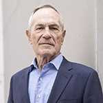 A man in a navy blue suit and light blue collared shirt stands and smiles