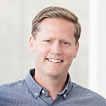 A man in a gray collared shirt stands and smiles