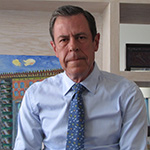 A man in a light blue collared shirt and a blue tie stands and smiles.