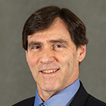 A man in a black blazer and blue collared shirt stands and smiles