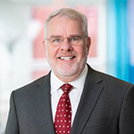 A man  in a black blazer, white collared shirt, and red tie stands and smiles