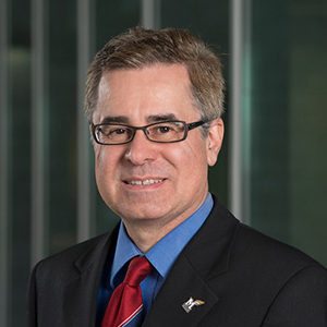 A man in a black suit, blue collared shirt, and red tie stands and smiles.