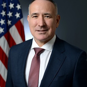 A man in a navy blue suit, white collared shirt, and maroon tie stands in front of the American flag and smiles.