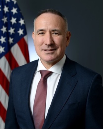 A man in a navy blue suit, white collared shirt, and maroon tie stands in front of the American flag and smiles.