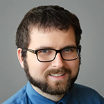 A man in glasses and a blue collared shirt stands and smiles