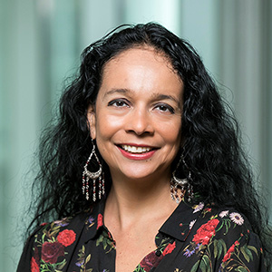 Professional headshot of a smiling woman with long curly black hair, wearing dangling earrings and a black floral blouse, posed against a softly blurred light-colored background.
