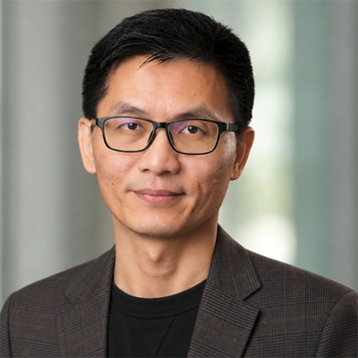 Professional headshot of a person wearing glasses and a dark blazer over a black shirt, facing the camera with a neutral expression against a softly blurred indoor background.