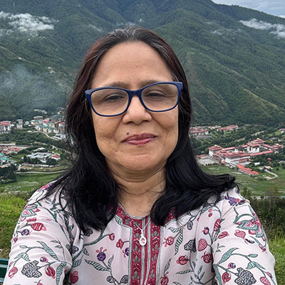 Portrait of a woman wearing blue glasses and a patterned blouse, smiling outdoors with green mountains and a valley town in the background.