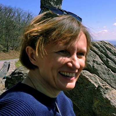Close-up of a smiling person outdoors near large rocks and a winding road, with short light brown hair and sunglasses resting on their head under a clear blue sky.