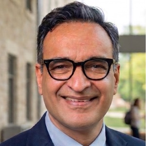 Portrait of a smiling man wearing glasses, a suit jacket, and a blue shirt, standing outdoors in front of a campus building.