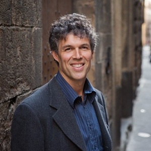 A smiling man with curly dark hair wearing a dark blazer and blue shirt, standing outdoors beside a stone wall.